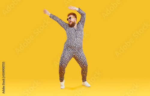 Cheerful funny eccentric fat man in good mood is dancing and fooling around on orange background. Overweight bearded man dressed in leopard print jumpsuit is dancing and fooling around. Full length.