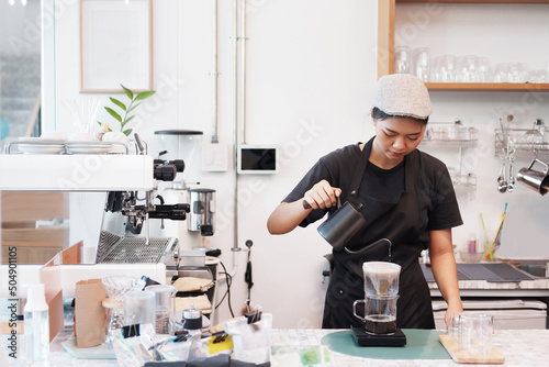 Smiling Asian Young barista woman is wearing apron and brewing coffee with machine for customers order in cafe and coffee shop. Start up small cafe business and technology Concept.