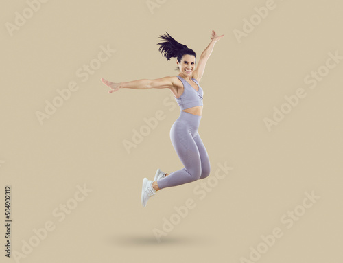 Happy excited cheerful joyful beautiful fit young woman in lilac crop top and leggings smiling and jumping against beige studio background. Sport, fitness, workout, energy, healthy lifestyle concept