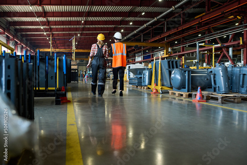 Rear view of two young female engineers in workwear moving along wide aisle in large workshop of indistrial plant or modern factory