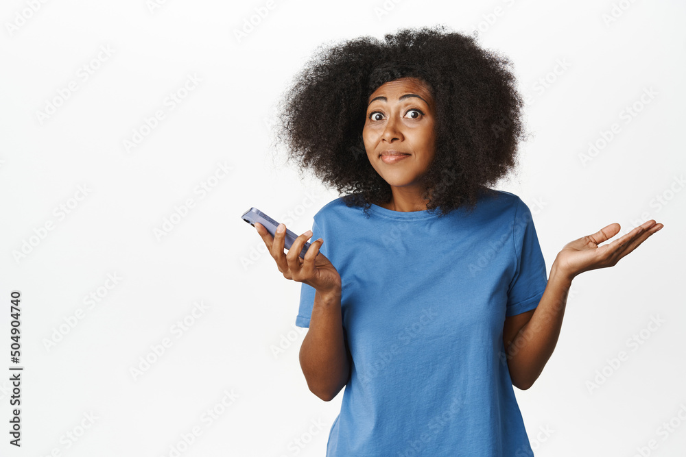 Dont know. Clueless african american woman shrugs shoulders with mobile phone and smiles, confused, puzzled to answer, stands in blue t-shirt over white background Dont know. Clueless african american woman shrugs shoulders with mobile phone and smiles, confused, puzzled to answer, stands in blue t-shirt over white background
