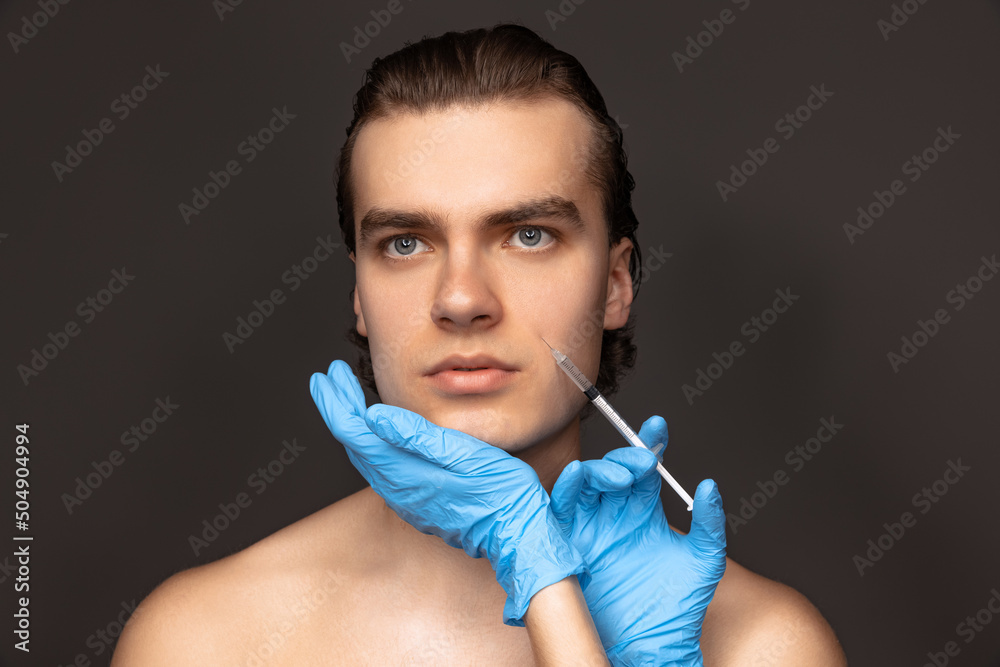 Close-up face of young man and female hands in blue gloves with syringe isolated on dark background. Cosmetic, filling surgery procedures. Anti-aging concept.