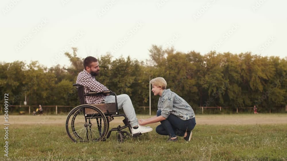 Loving couples ties his shoelaces to disabled person man sitting in wheelchair on grass in park, side view. Concept of care and love Loving couples ties his shoelaces to disabled person man sitting in wheelchair on grass in park, side view. Concept of care and love