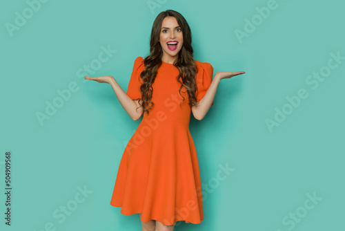 Fotografia Happy young woman in orange mini dress is posing with hands raised and shouting
