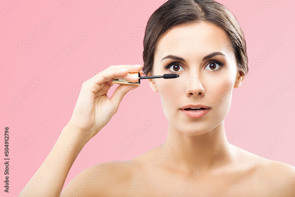 Portrait of beautiful woman holding in hand, putting mascara on long thick eye lashes with brush, isolated over rose pink background. Brunette girl at beauty treatment, visage, makeup studio concept.