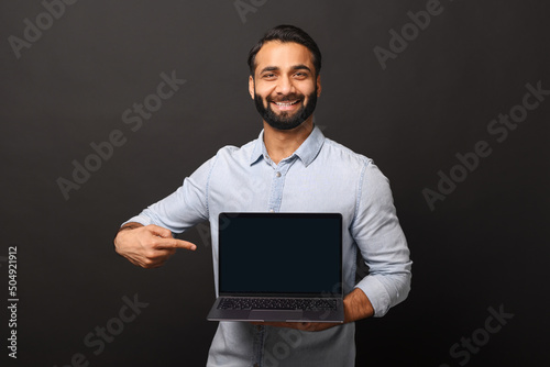 Happy Indian man holding laptop with empty screen and points finger at it, hispanic male employee presenting new computer app, promotion and advertise concept
