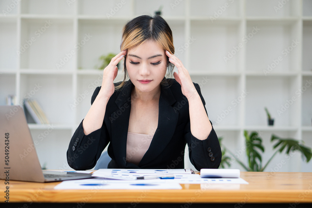Asian woman with headache working on a laptop and analyzing financial charts on the table Asian woman with headache working on a laptop and analyzing financial charts on the table