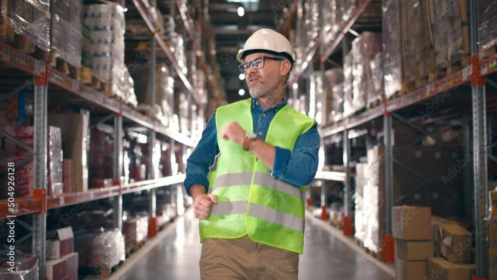 Happy positive Caucasian middle-aged man in helmet and eyeglasses walking in rows with many shelves with goods in boxes and dancing in good mood. Construction, factory, logistics center, work concept Happy positive Caucasian middle-aged man in helmet and eyeglasses walking in rows with many shelves with goods in boxes and dancing in good mood. Construction, factory, logistics center, work concept