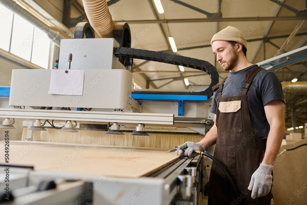 Young carpenter in apron working with wooden boards at modern machine at furniture factory Young carpenter in apron working with wooden boards at modern machine at furniture factory