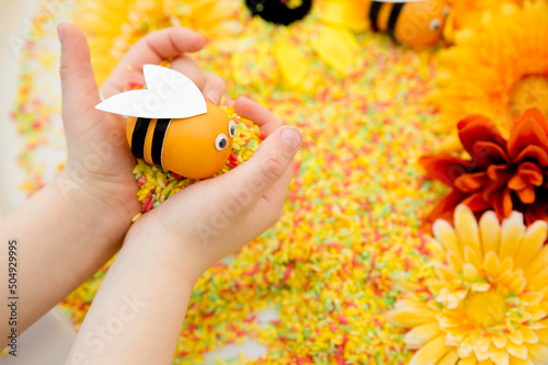 Summer in the meadow. Child plays with bee toy on colorful rice filled with artificial blooming flowers. Symbol of beekeeping, pollination and Bee collecting pollen from daisy. Sensory therapy.