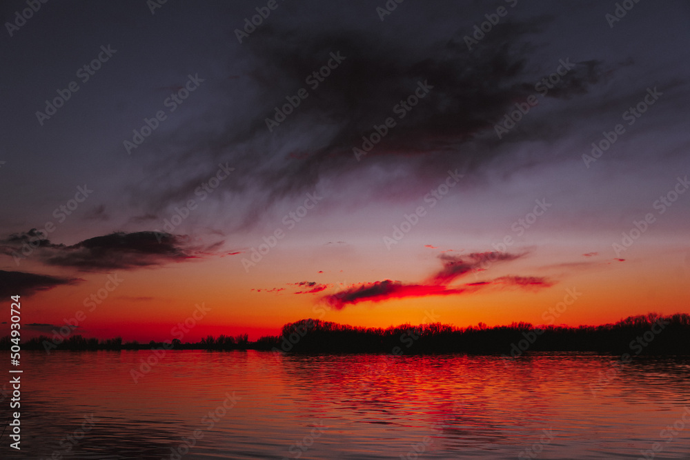 Orange, yellow, pink, blue and purple sunset sky  with dark colorful clouds with black trees reflection on calm river water	