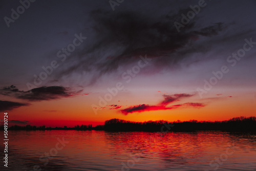 Orange, yellow, pink, blue and purple sunset sky  with dark colorful clouds with black trees reflection on calm river water  © kapichka