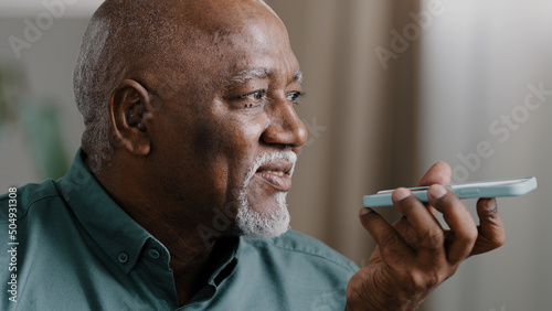 Mature grey-haired 60s biracial man with beard african american senior old male at home hold smartphone speak to phone microphone speaker audio voice message using speakerphone talking mobile app