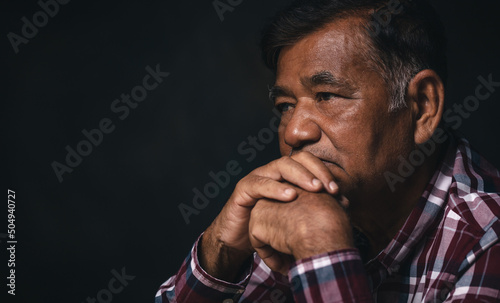 Elderly man bowed his head sit praying to God on black background at home. An old man thinking alone.