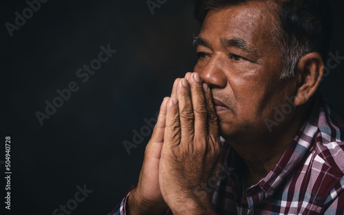 Elderly man praying to God on black background at home. © Pituk