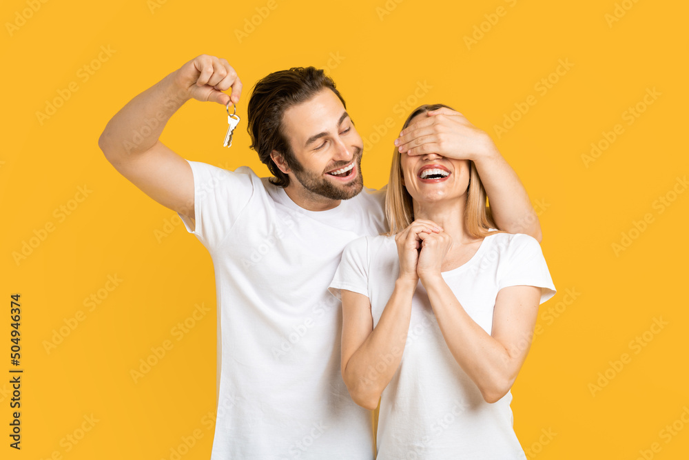 Smiling young european guy in white t-shirt closes eyes to his wife and holds keys, isolated on yellow background Smiling young european guy in white t-shirt closes eyes to his wife and holds keys, isolated on yellow background