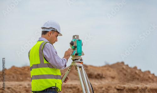 Surveyor engineer wearing safety uniform and helmet with equipment theodolite to measurement positioning on the construction site of the road with construct machinery background