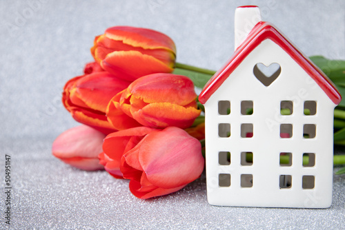 A white house with a red roof and tulips on a silver background.The theme of love and celebration.The concept of delivery and mortgage. Birthday or Valentine's Day
