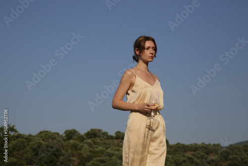 Outdoor portrait of woman in beige silk outfit holding a glass of white wine. Idyllic summer vacation concept. 