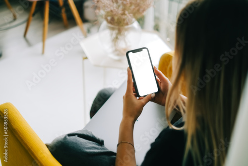 Woman holding mobile phone with white screen mock up, resting on a sofa in living room at home.
