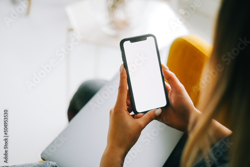 Woman holding mobile phone with white screen mock up, resting on a sofa in living room at home.