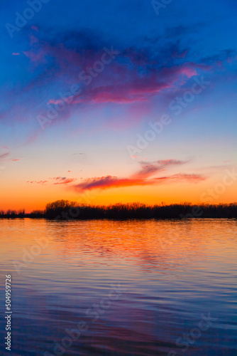 Orange, yellow and purple sunset on river with dark colorful clouds in sky with trees reflection in water