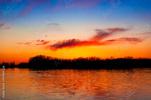 Orange, yellow and purple sunset on river with dark colorful clouds in sky with trees reflection in water