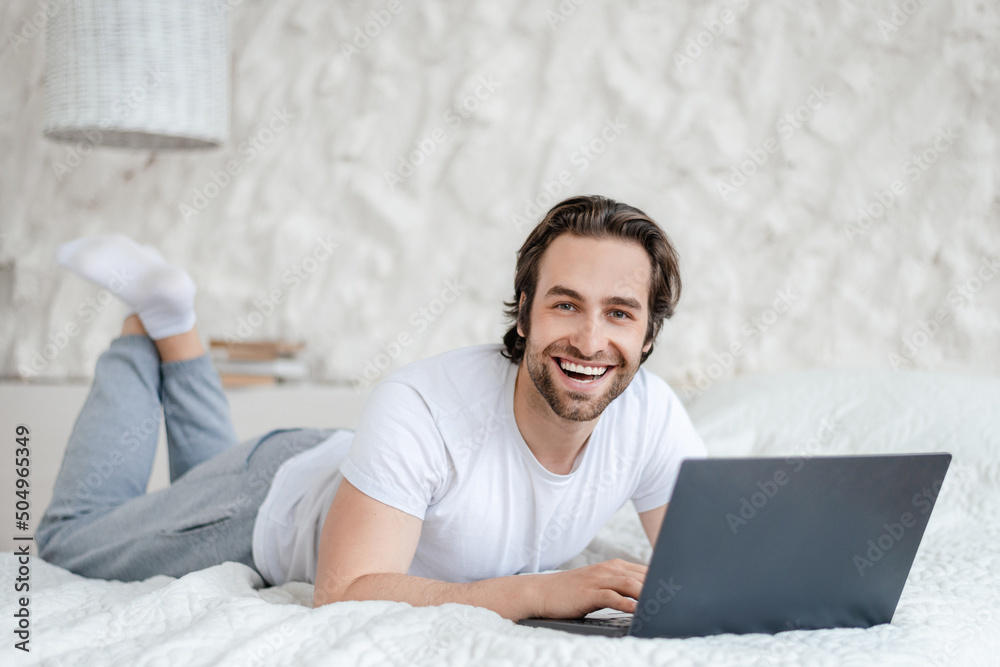 Glad young caucasian man with stubble lies on bed with computer, surfing in internet in light bedroom interior Glad young caucasian man with stubble lies on bed with computer, surfing in internet in light bedroom interior