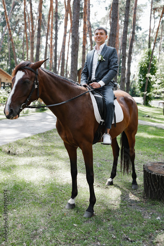 The groom in a blue suit on a brown horse in the woods, fooling around.