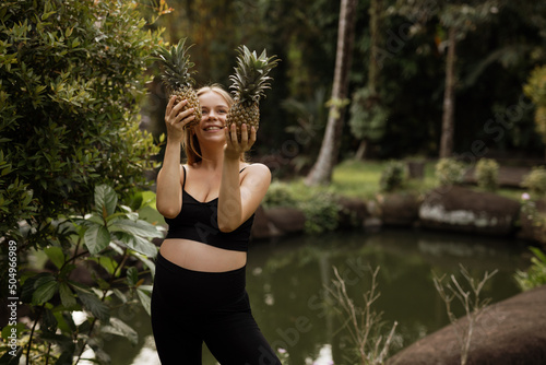 Pregnant girl stands in a garden near a pond and holds fruits in her hands. Girl in black clothes with pineapples