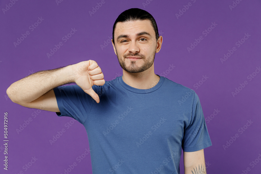 Obraz premium Young displeased dissatisfied sad disappointed man 20s wear basic blue t-shirt showing thumb down dislike gesture isolated on plain purple color background studio portrait. People lifestyle concept