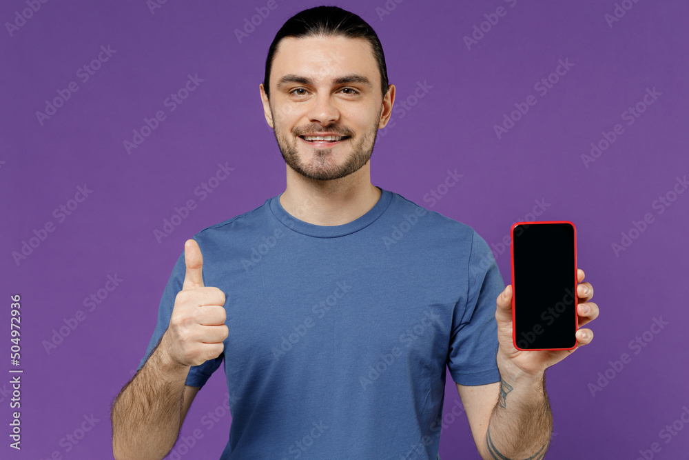 Young happy fun man 20s wearing basic blue t-shirt hold in hand use mobile cell phone with blank screen workspace area show thumb up gesture isolated on plain purple color background studio portrait Young happy fun man 20s wearing basic blue t-shirt hold in hand use mobile cell phone with blank screen workspace area show thumb up gesture isolated on plain purple color background studio portrait