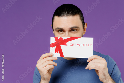 Young happy man 20s wearing basic blue t-shirt hold cover mouth with gift certificate coupon voucher card for store isolated on plain purple color background studio portrait. People lifestyle concept.
