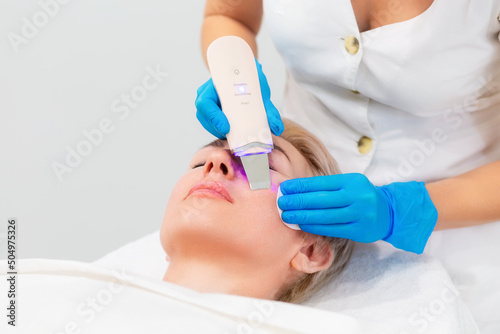 A professional cosmetologist in blue medical gloves does an ultrasound facial cleansing for an adult Caucasian woman. Cleansing device on the client s cheek. Hardware anti-aging cosmetology