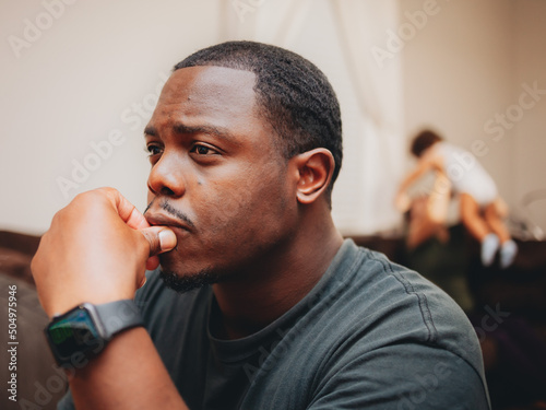 close up of African American or black man at home sitting looking depressed and worried while wife and kids play in the background