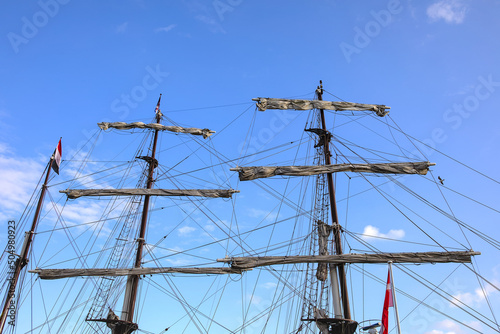 Masts, rigging and sails of an old historical sailing ship in a harbor against a blue sky with few clouds