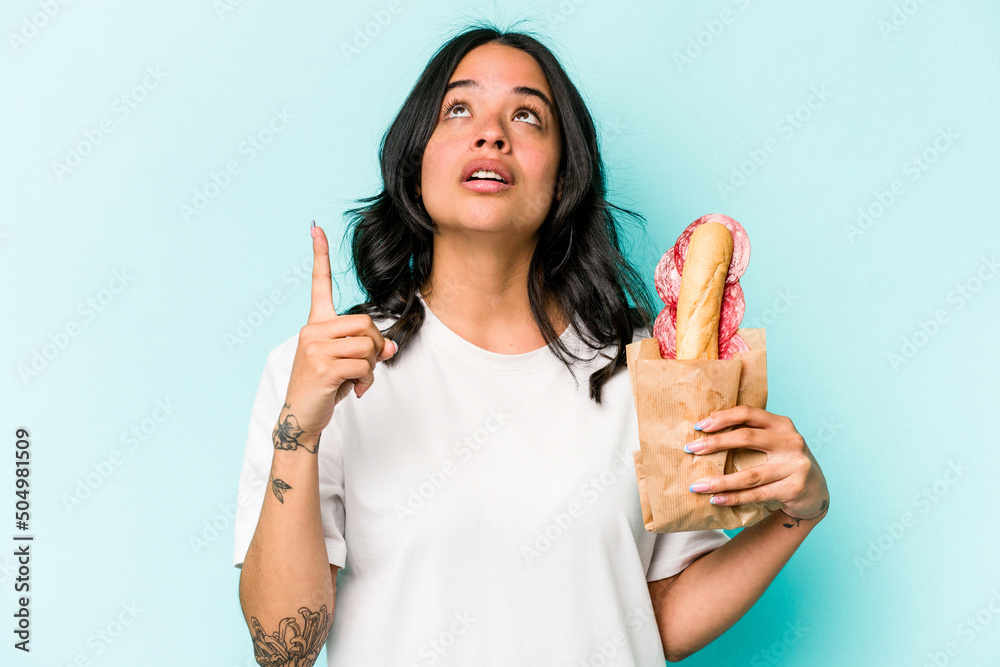 Young hispanic woman eating a sandwich isolated on blue background pointing upside with opened mouth. Young hispanic woman eating a sandwich isolated on blue background pointing upside with opened mouth.