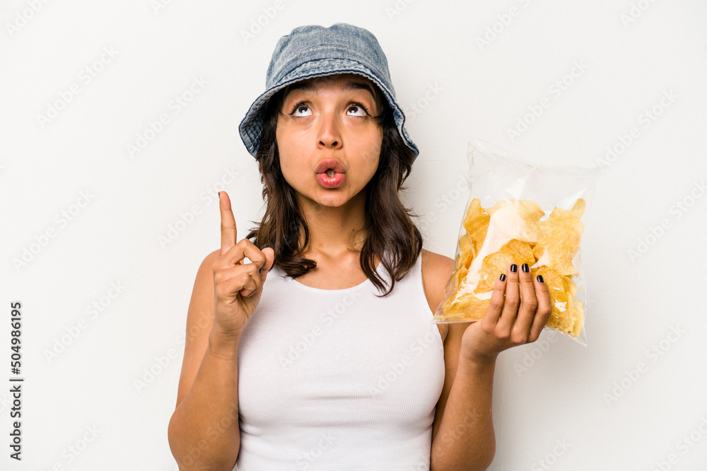 Young hispanic woman holding a bag of chips isolated on white background pointing upside with opened mouth. Young hispanic woman holding a bag of chips isolated on white background pointing upside with opened mouth.