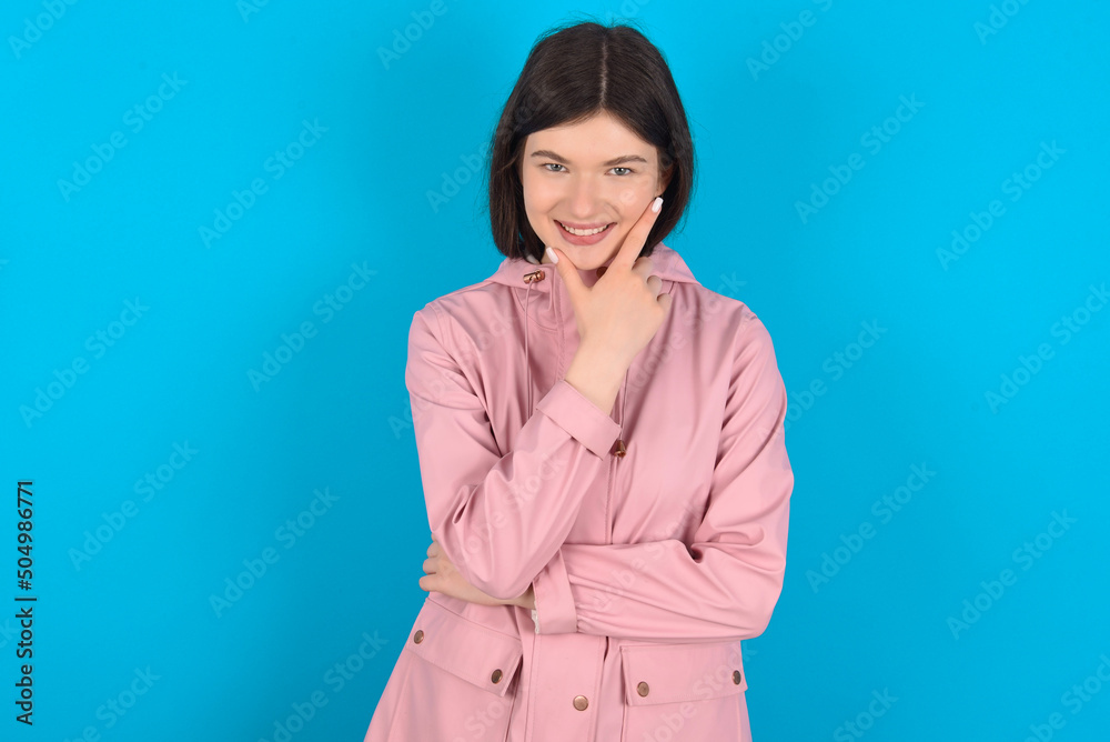young beautiful Caucasian woman wearing pink raincoat over blue wall looking confident at the camera smiling with crossed arms and hand raised on chin. Thinking positive. young beautiful Caucasian woman wearing pink raincoat over blue wall looking confident at the camera smiling with crossed arms and hand raised on chin. Thinking positive.