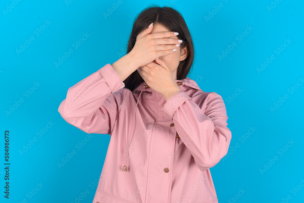 young beautiful Caucasian woman wearing pink raincoat over blue wall Covering eyes and mouth with hands, surprised and shocked. Hiding emotions. young beautiful Caucasian woman wearing pink raincoat over blue wall Covering eyes and mouth with hands, surprised and shocked. Hiding emotions.
