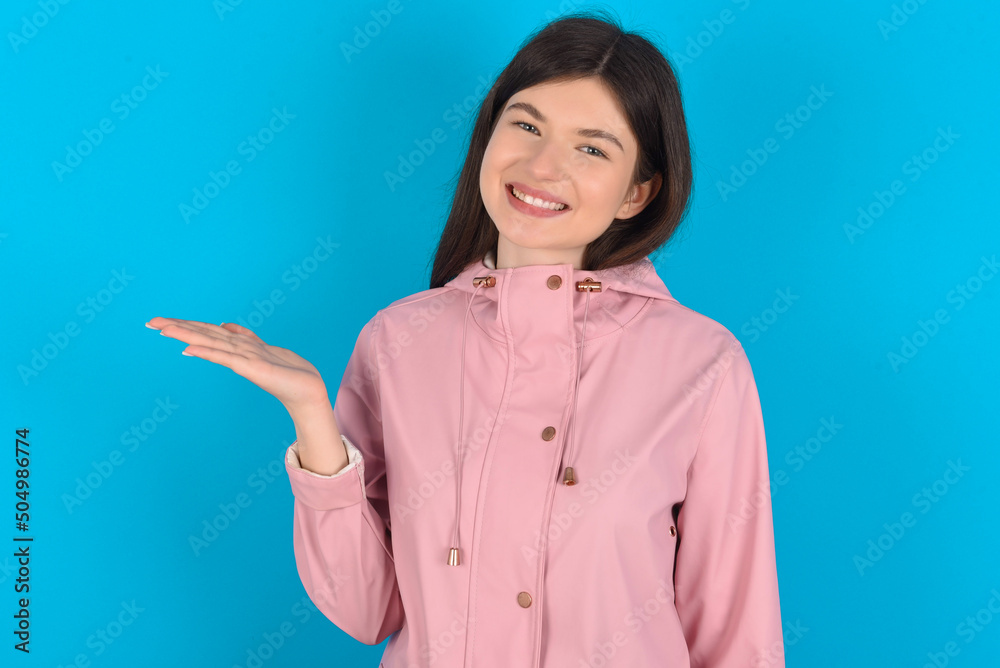 young beautiful Caucasian woman wearing pink raincoat over blue wall smiling cheerful presenting and pointing with palm of hand looking at the camera. young beautiful Caucasian woman wearing pink raincoat over blue wall smiling cheerful presenting and pointing with palm of hand looking at the camera.