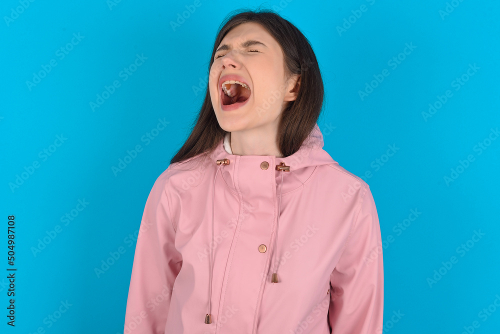 young beautiful Caucasian woman wearing pink raincoat over blue wall angry and mad screaming frustrated and furious, shouting with anger. Rage and aggressive concept. young beautiful Caucasian woman wearing pink raincoat over blue wall angry and mad screaming frustrated and furious, shouting with anger. Rage and aggressive concept.