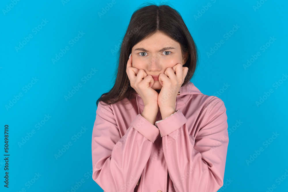 Fearful young beautiful Caucasian woman wearing pink raincoat over blue wall keeps hands near mouth, feels frightened and scared, has a phobia, Shock and frighted concept. Fearful young beautiful Caucasian woman wearing pink raincoat over blue wall keeps hands near mouth, feels frightened and scared, has a phobia, Shock and frighted concept.