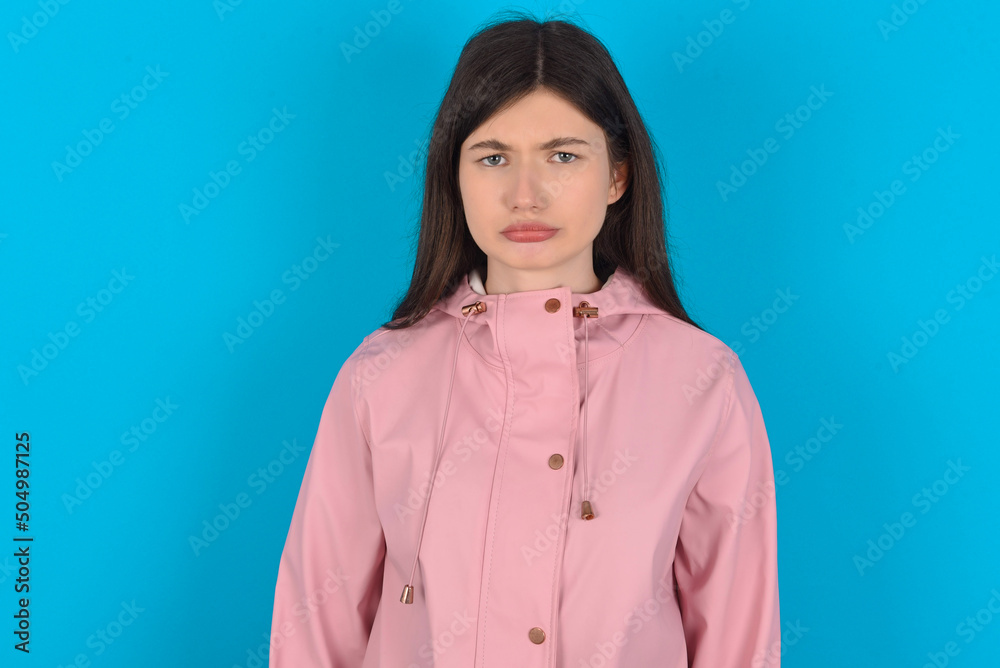 Displeased upset young beautiful Caucasian woman wearing pink raincoat over blue wall frowns face as going to cry, being discontent and unhappy as can't achieve goals, Disappointed model has troubles Displeased upset young beautiful Caucasian woman wearing pink raincoat over blue wall frowns face as going to cry, being discontent and unhappy as can't achieve goals, Disappointed model has troubles
