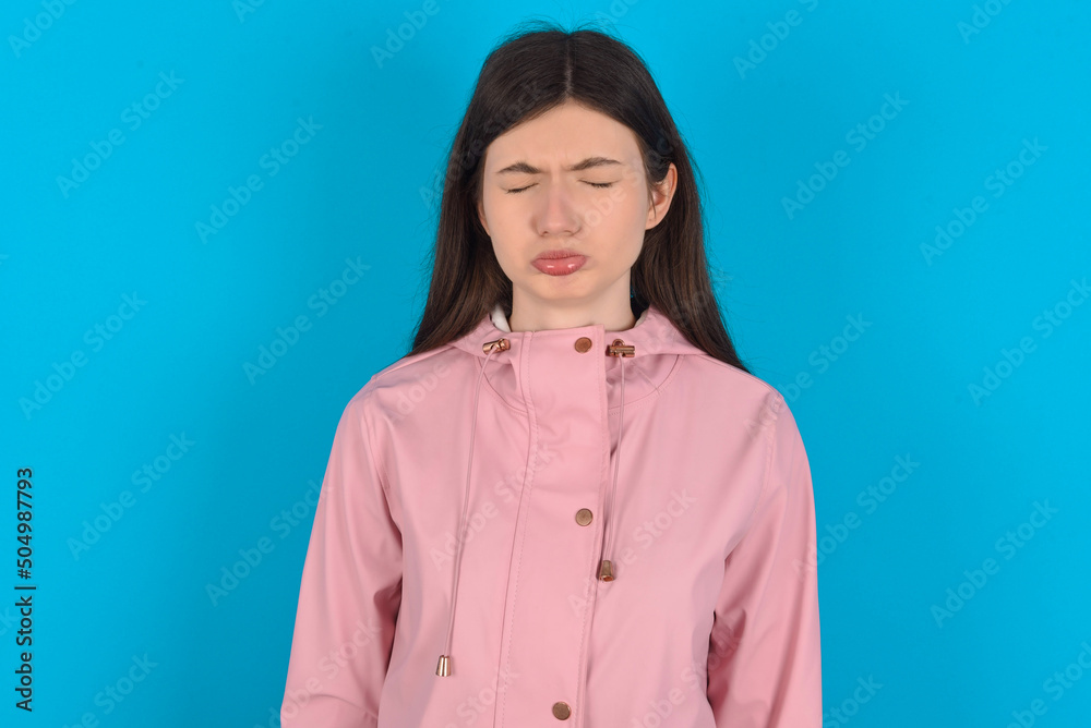 Dismal gloomy rejected young beautiful Caucasian woman wearing pink raincoat over blue wall has problems and difficulties, curves lower lip and closes eyes in despair, being in depression Dismal gloomy rejected young beautiful Caucasian woman wearing pink raincoat over blue wall has problems and difficulties, curves lower lip and closes eyes in despair, being in depression