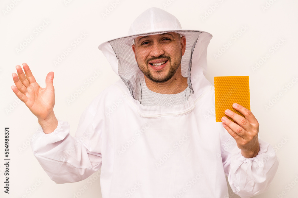 Hispanic beekeeper man holding honey isolated on white background receiving a pleasant surprise, excited and raising hands. Hispanic beekeeper man holding honey isolated on white background receiving a pleasant surprise, excited and raising hands.