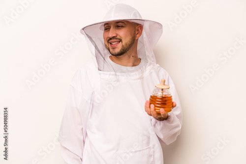 Hispanic beekeeper man holding honey isolated on white background looks aside smiling, cheerful and pleasant.