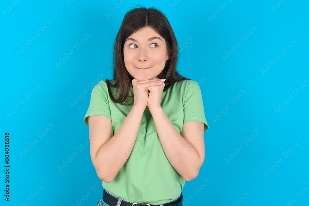 Curious young beautiful Caucasian woman wearing green T-shirt over blue wall keeps hands under chin bites lips and looks with interest aside. Curious young beautiful Caucasian woman wearing green T-shirt over blue wall keeps hands under chin bites lips and looks with interest aside.