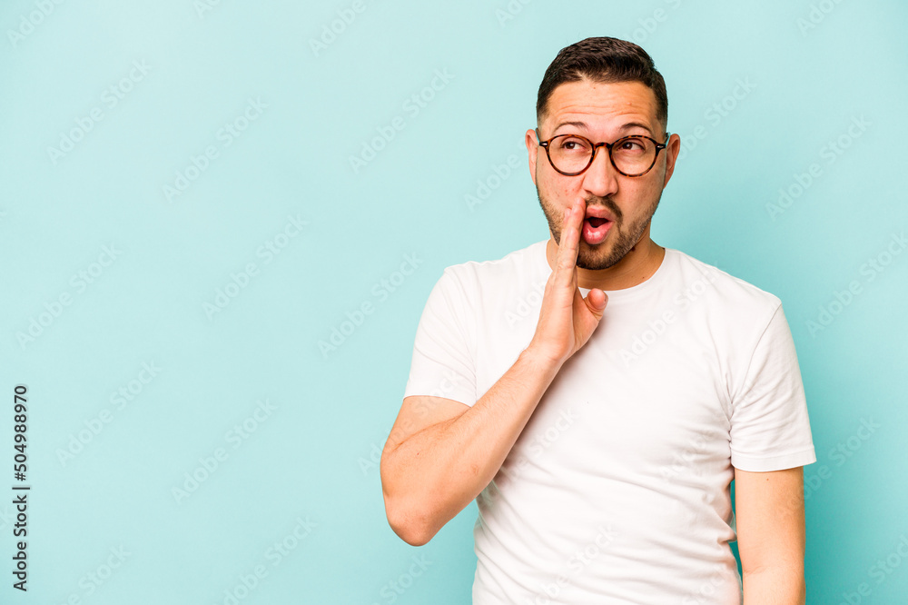 Young hispanic man isolated on blue background is saying a secret hot braking news and looking aside Young hispanic man isolated on blue background is saying a secret hot braking news and looking aside
