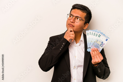 Young hispanic business man holding banknotes isolated on white background looking sideways with doubtful and skeptical expression.